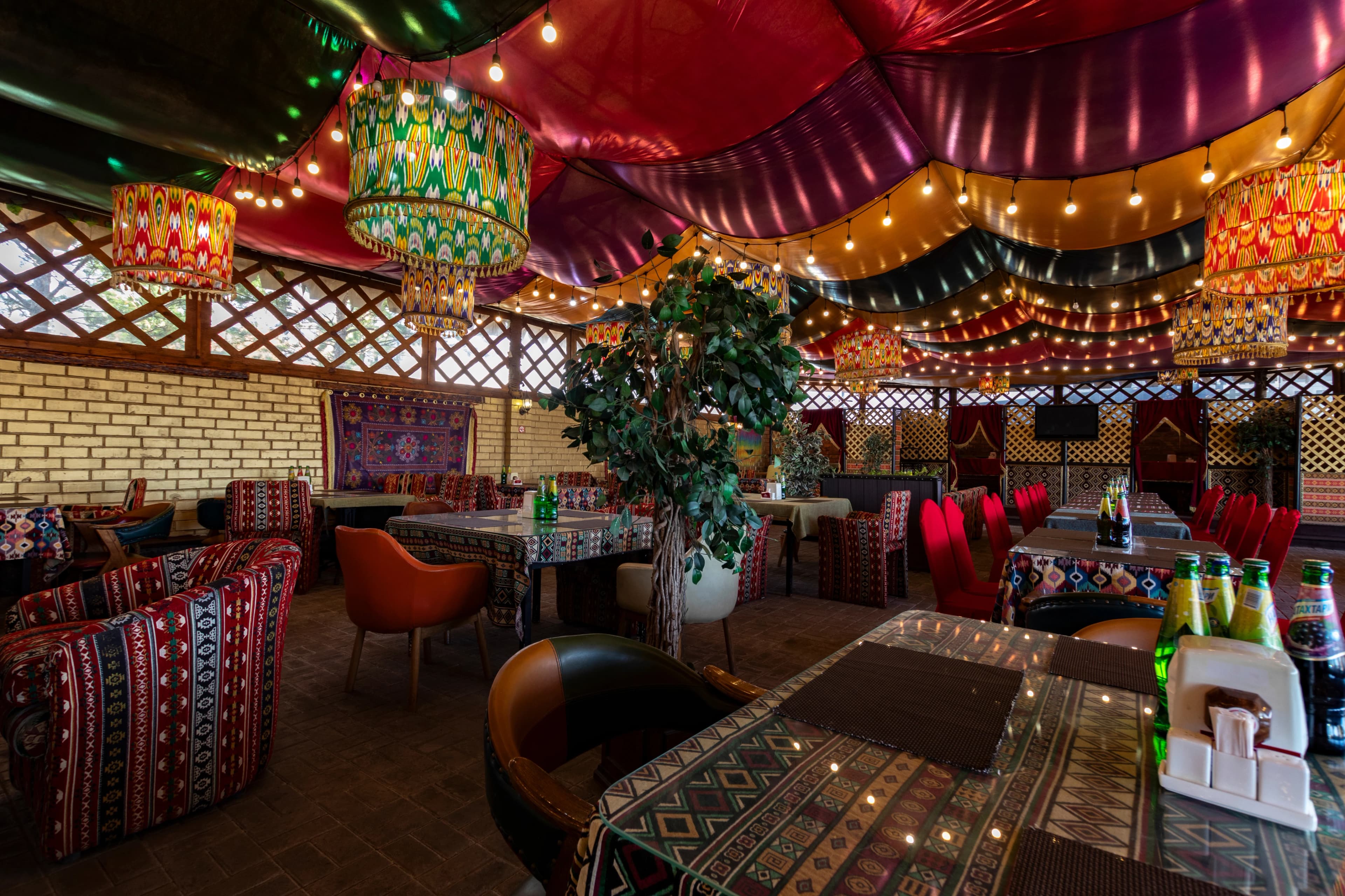 Панорамный вид на пустой просторный зал ресторана с современным интерьером: ряды столов со светлыми столешницами и стульями жёлтого и тёмно‑синего цвета, над ними висят цилиндрические подвесные светильники, на столах расставлены бутылки, бокалы и приборы, потолок украшен извилистыми красно‑жёлтыми конструкциями, а в глубине видны арочные проёмы и барная стойка.