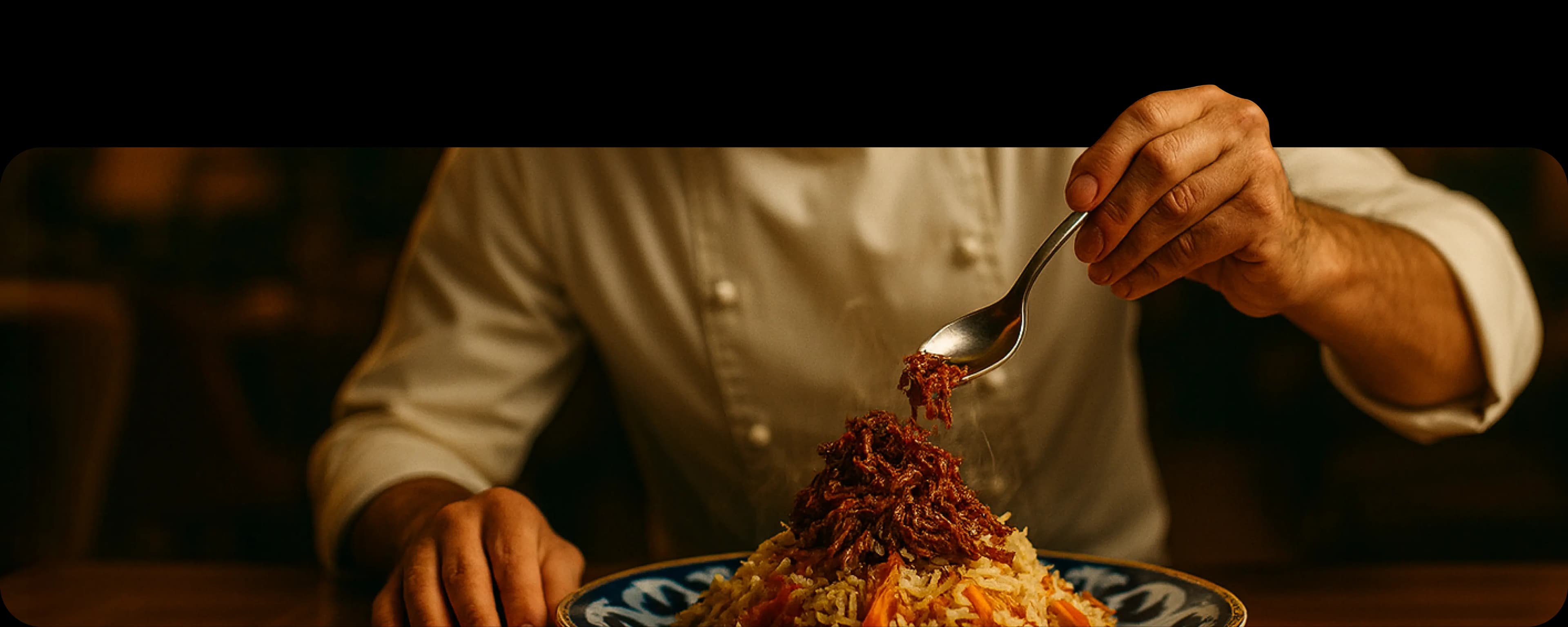 Рука держит ложку, из которой стекает соус на изысканно сервированное блюдо на тарелке, на фоне приглушённого интерьера и человека в белой рубашке.