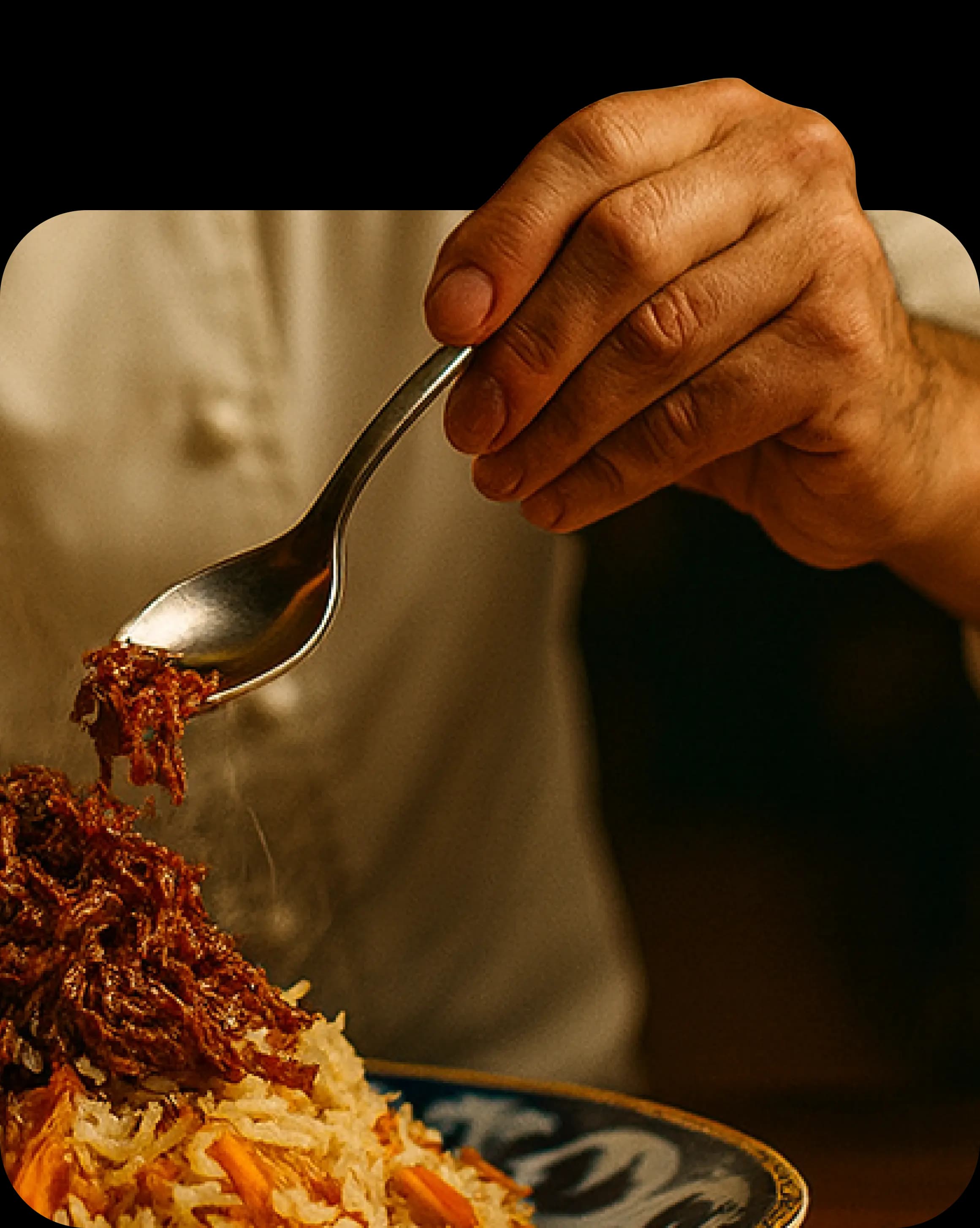 Рука держит ложку, из которой стекает соус на изысканно сервированное блюдо на тарелке, на фоне приглушённого интерьера и человека в белой рубашке.