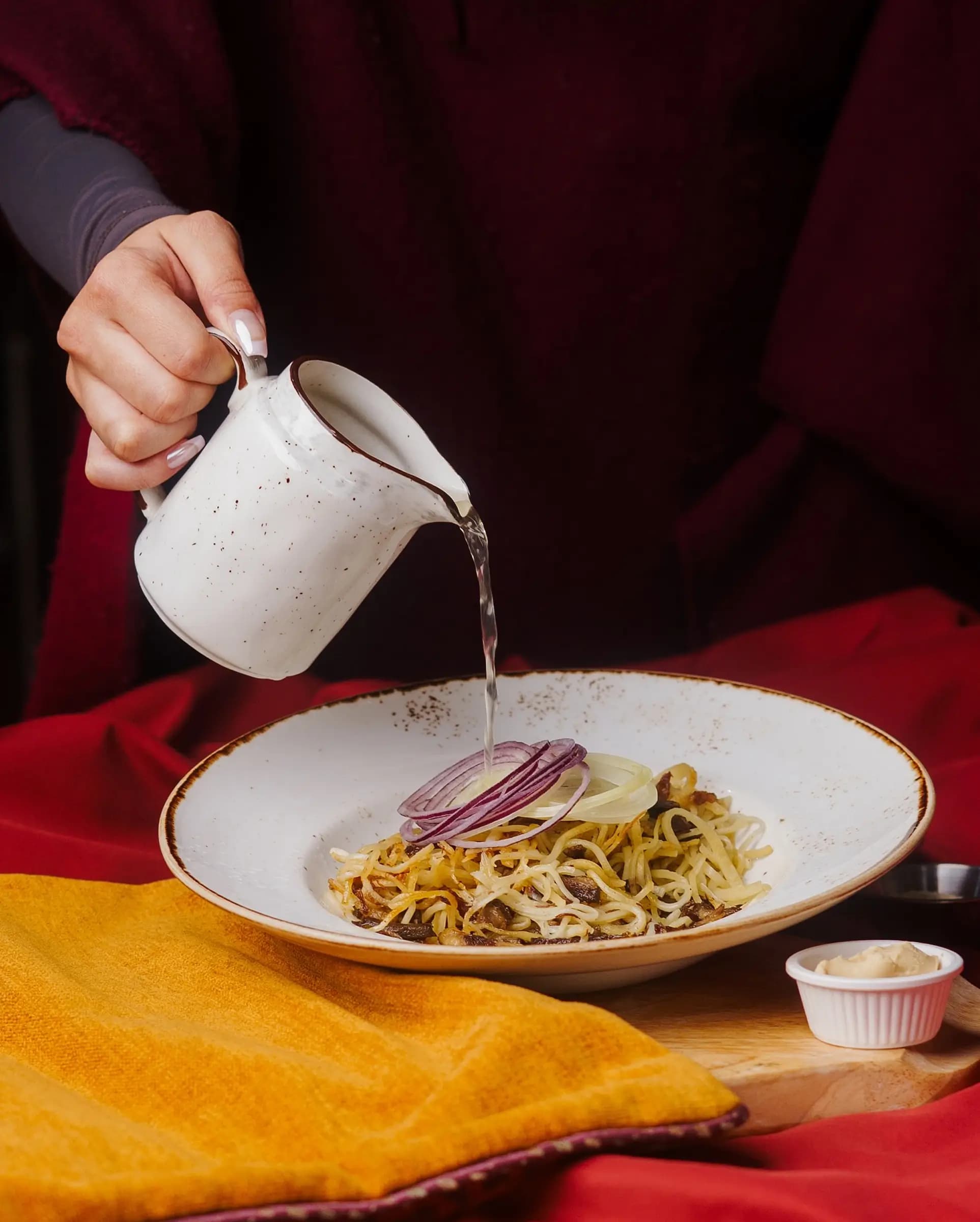 Рука наливает белый соус на тарелку с желтыми спагетти, украшенными луком и грибами, на красном фоне с желтым шарфом.