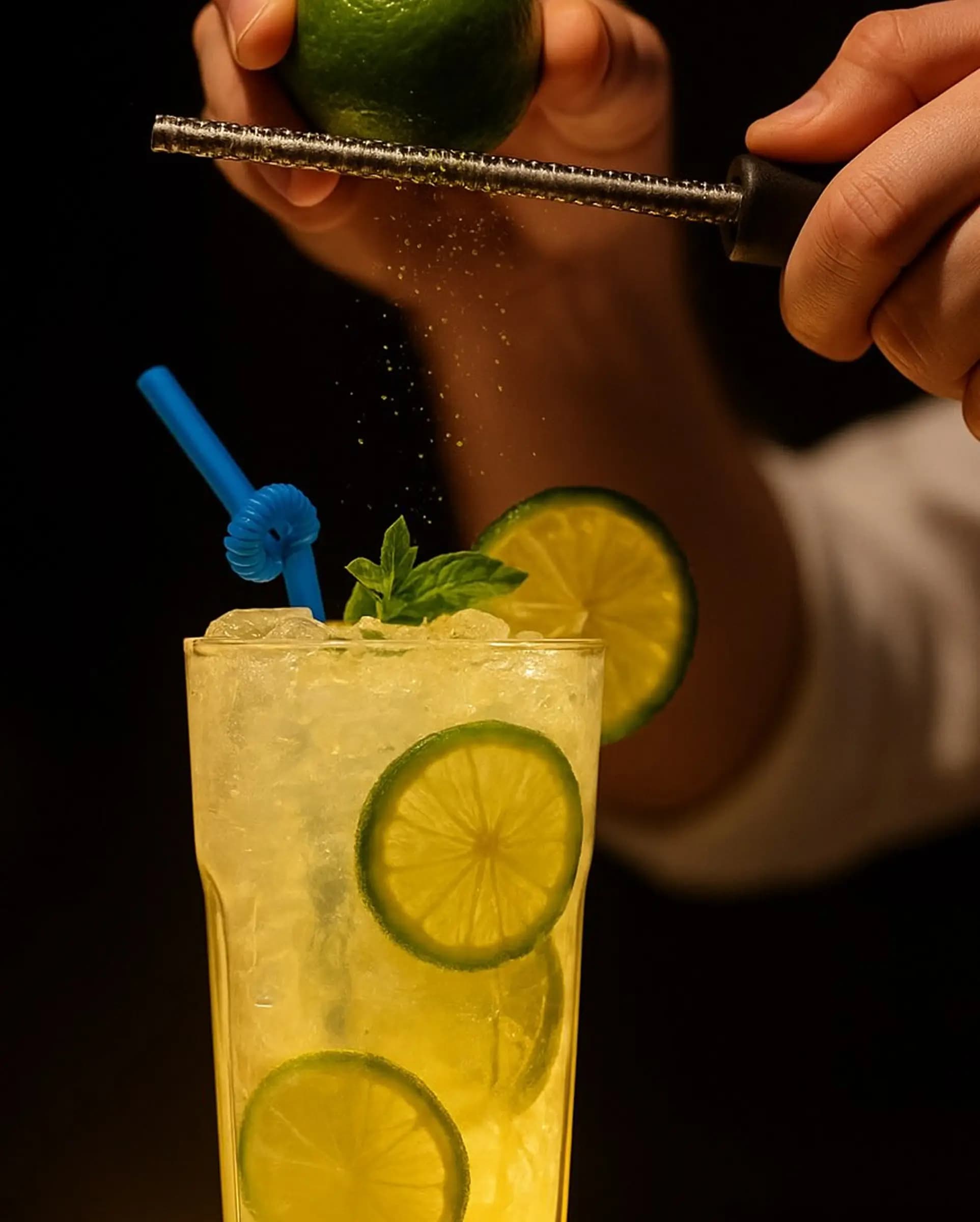 Бармен зестит лайм над коктейлем с льдом, мятой и ломтиками лайма в высоком стакане, с синей соломинкой.