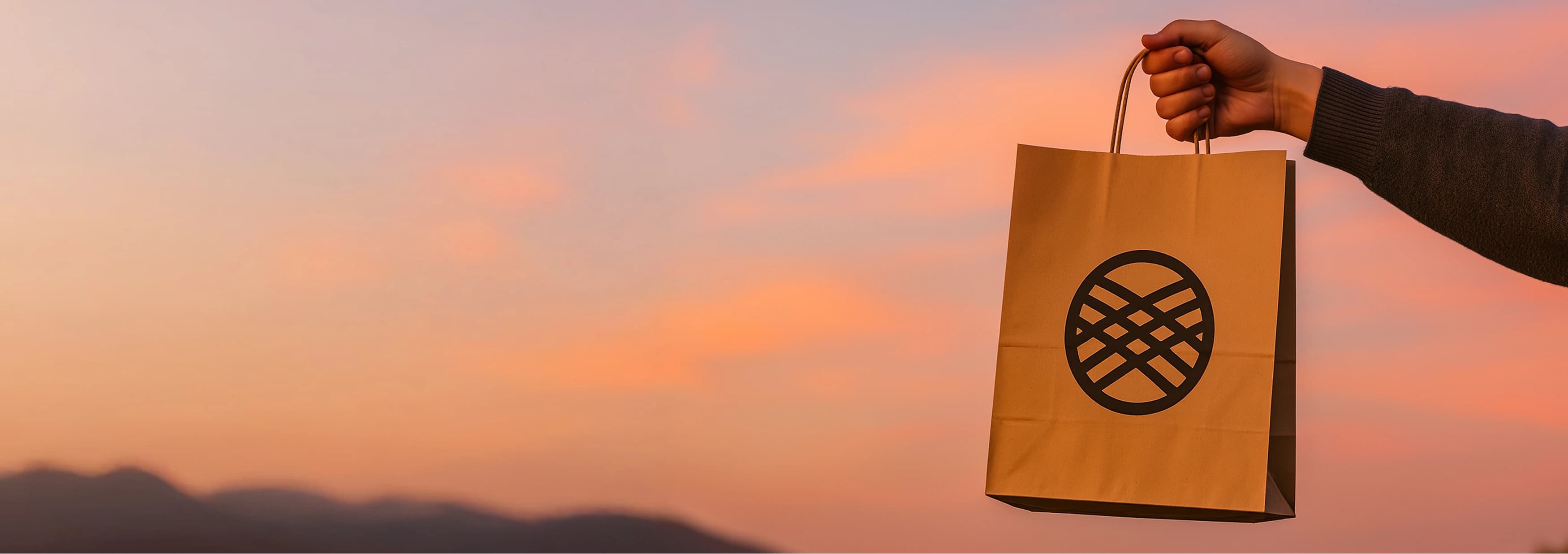 Рука держит бумажный пакет с логотипом на фоне закатного неба.